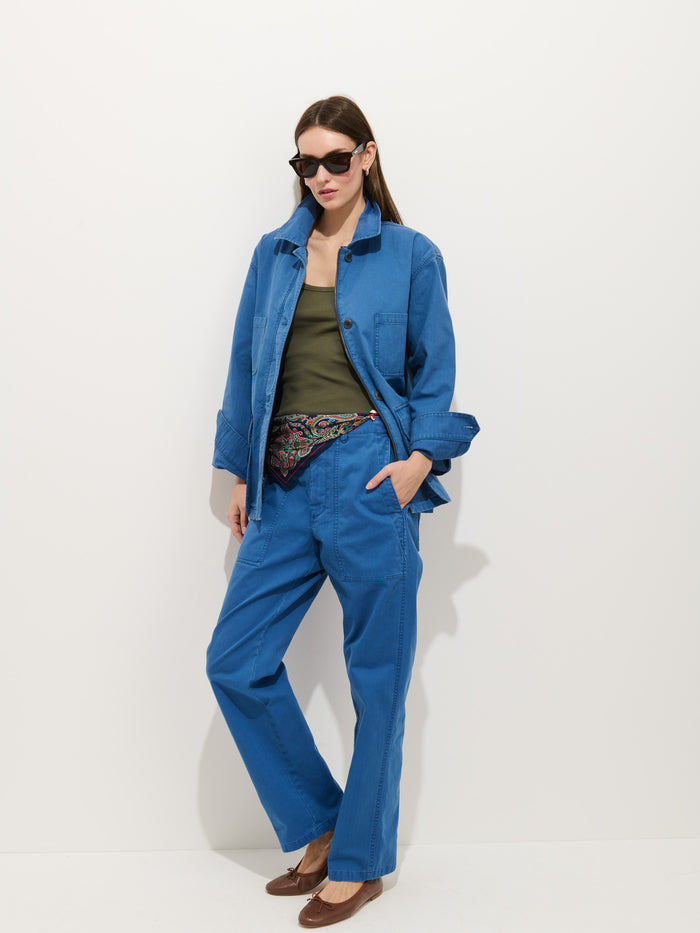 A woman stands against a white wall in the Painter Jacket In Herringbone, matching pants, an olive green top, brown flats, black sunglasses, and a patterned scarf tucked into her waistband.