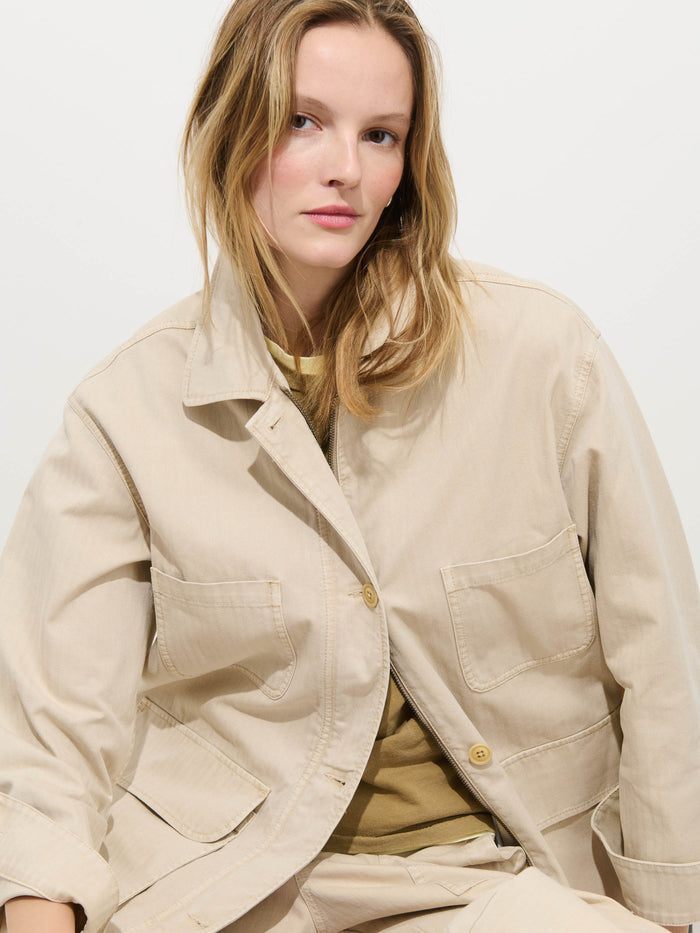 A woman with light brown hair models the oversized Painter Jacket In Herringbone over a tan shirt, sitting against a plain white background and looking at the camera with a neutral expression.