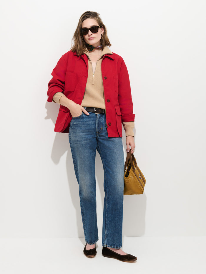A woman wearing sunglasses, the oversized fit Painter Jacket In Herringbone in red cotton, a beige sweater, blue jeans, and brown flats stands against a white background, holding a mustard yellow handbag and posing with one hand in her pocket.