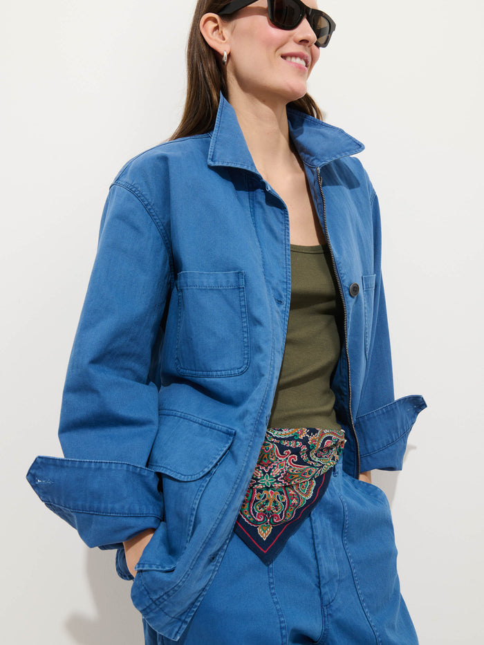 A person wearing the Painter Jacket In Herringbone, blue herringbone pants, an olive green tank top, dark sunglasses, and a colorful patterned scarf tucked into their waistband stands against a plain light background, smiling.