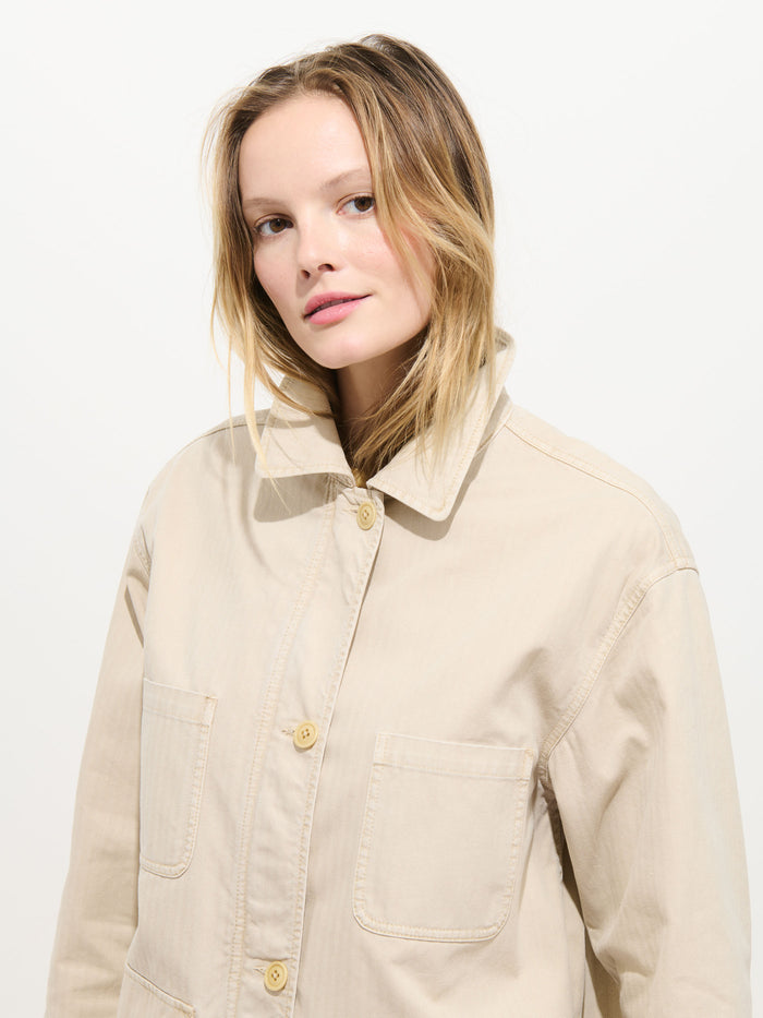A person with light skin and blonde hair wears the Painter Jacket In Herringbone, an oversized beige cotton jacket with large front pockets, and stands against a white background looking slightly to the side.