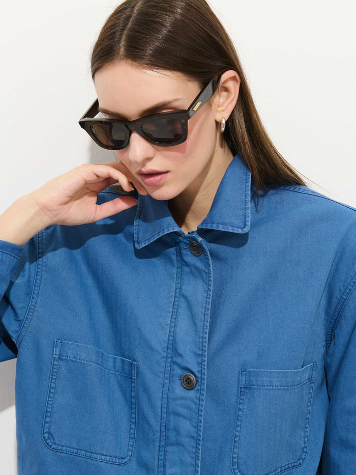 A woman with straight brown hair wears dark sunglasses, silver hoop earrings, and the Painter Jacket In Herringbone—an oversized blue cotton style with front pockets—while posing against a plain white background.