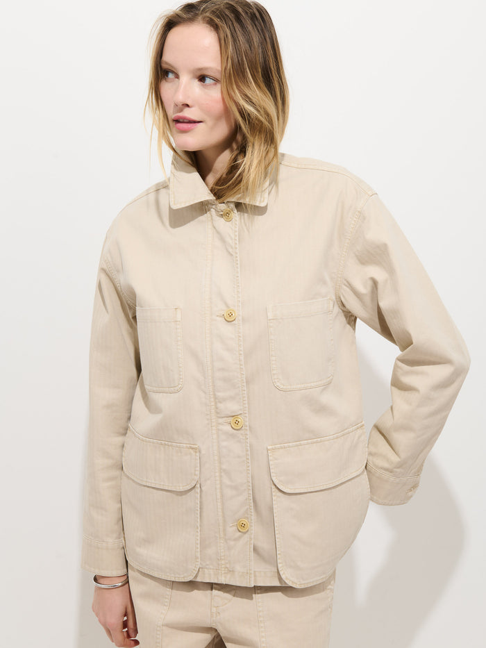 A woman with light hair wears the Painter Jacket In Herringbone, an oversized beige cotton utility jacket with large front pockets and matching pants. She stands against a white background, looking to the side with one hand on her hip.