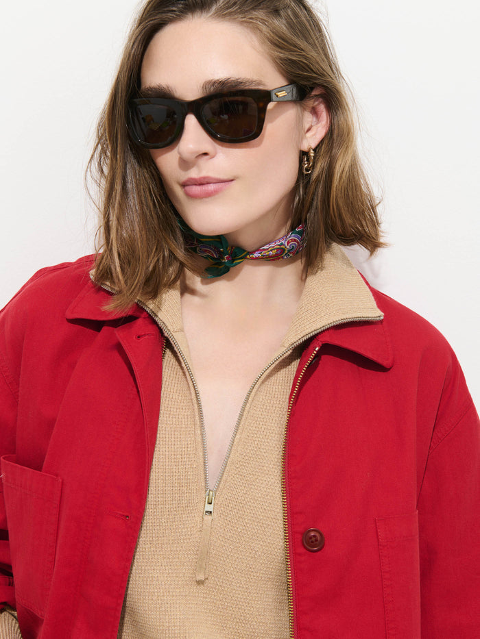 A woman wearing dark sunglasses, a Painter Jacket In Herringbone in an oversized red cotton fit, a beige zip-up top, and a colorful scarf around her neck stands against a plain white background.