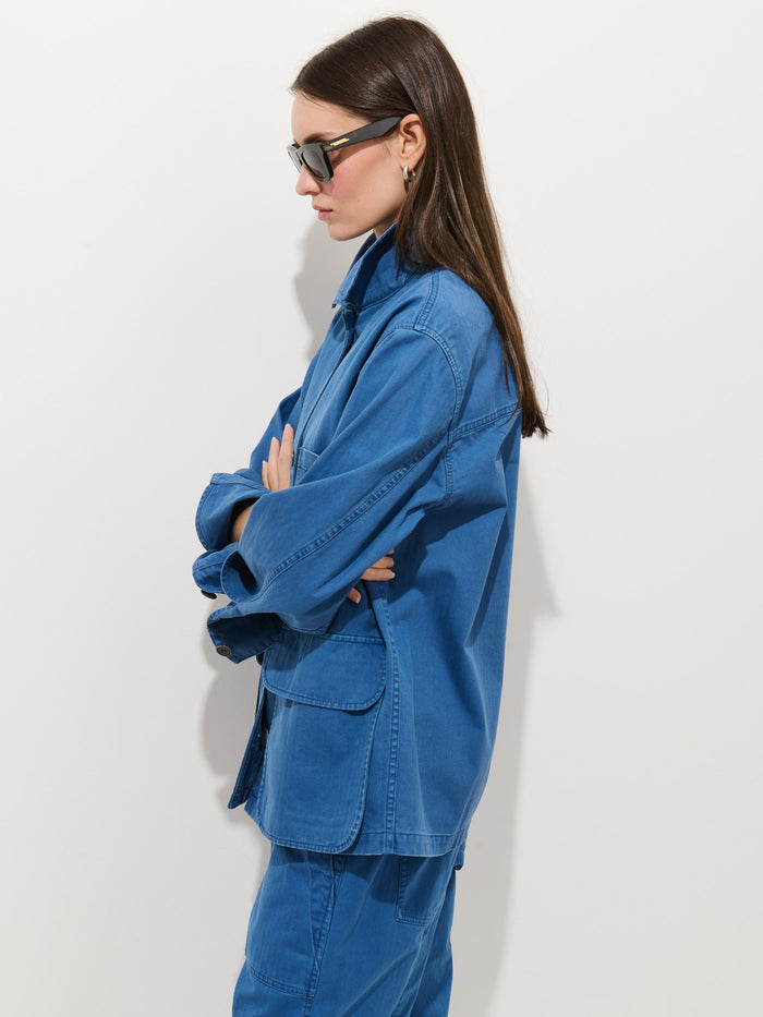 Woman with long brown hair wearing sunglasses and the Painter Jacket In Herringbone, styled oversized, with matching blue pants, standing in profile with arms crossed against a plain white background.