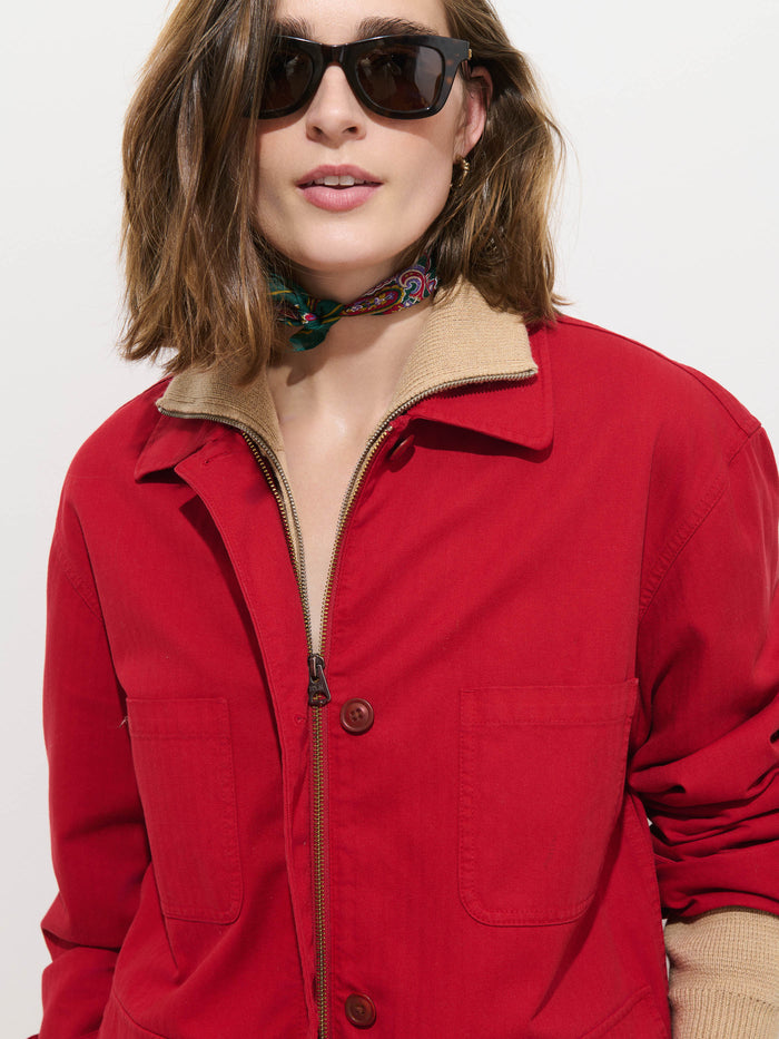 Woman wearing an oversized fit Painter Jacket In Herringbone in red cotton, paired with a beige top, sunglasses, and a colorful scarf around her neck, standing against a plain white background.