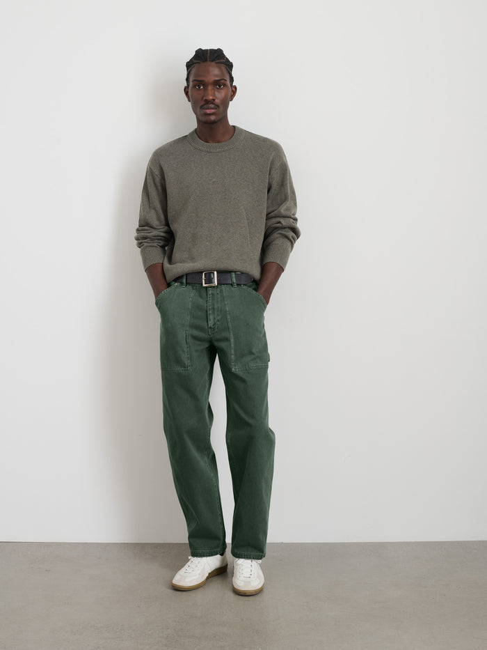 A man stands against a white wall, wearing the Painter Pant In Recycled Denim with a grey sweater, black belt, and white sneakers. He has his hands in his pockets and looks directly at the camera.