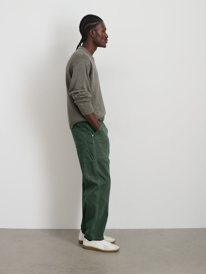 A person with braided hair stands in profile against a plain white wall, wearing a gray long-sleeve shirt, Painter Pant In Recycled Denim in green, and white sneakers.