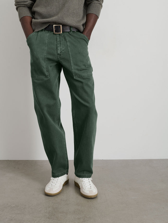 A person stands against a plain wall, wearing a grey sweater, Painter Pant In Recycled Denim in green, a black belt, and white sneakers. Their hands are tucked in their pockets and their face is not visible.