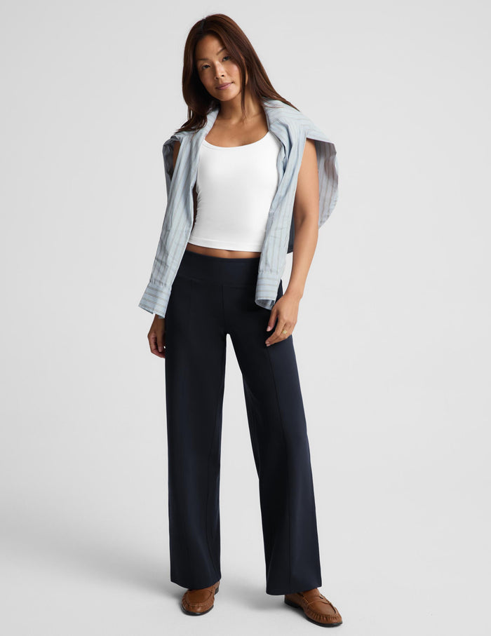 A woman modeling a pair of dark navy wide-leg trousers, styled with a white ribbed tank top, a light blue striped button-down shirt draped off her shoulders, and brown suede loafers, against a light gray studio background.