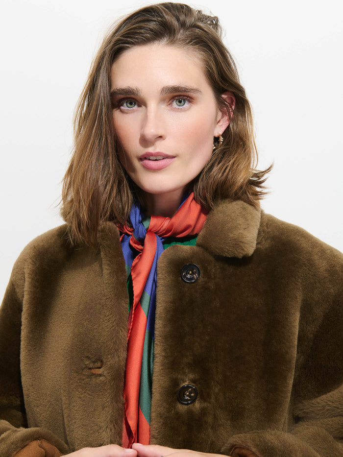 A woman with shoulder-length brown hair wears a brown faux fur coat and the Pop Art Silk Scarf, standing against a plain white background.