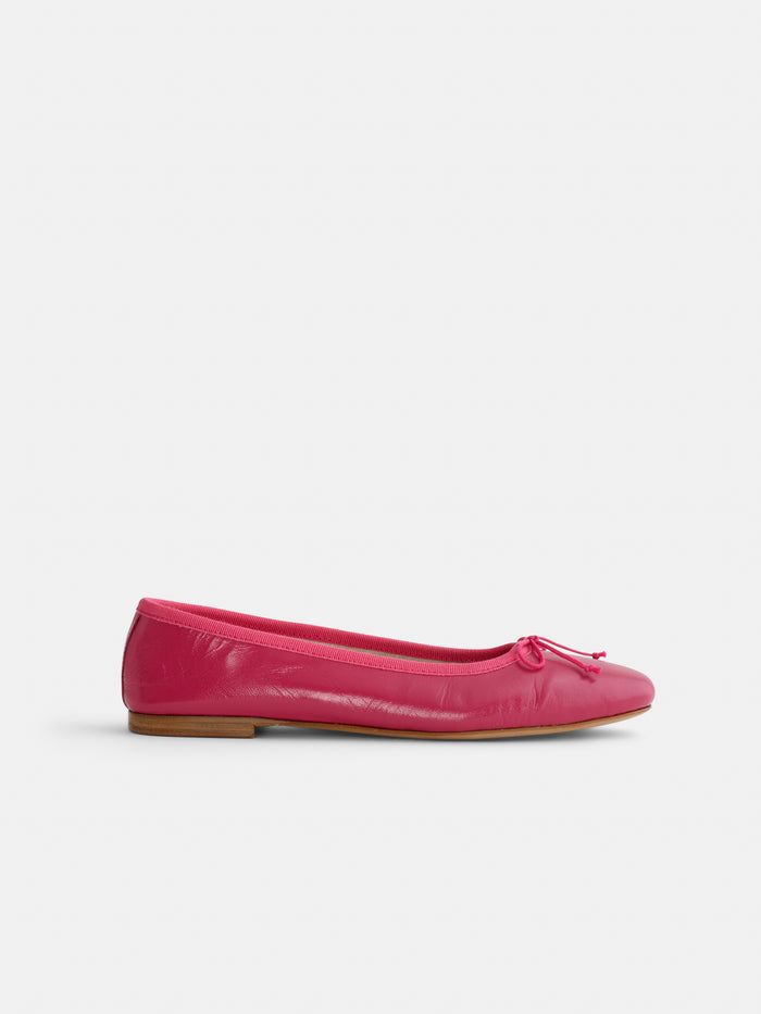 The Prince Street Ballet Flat is a bright pink shoe with a small bow on the rounded toe, low wooden heel, and shiny finish. Handmade in Florence, it's shown against a plain white background.