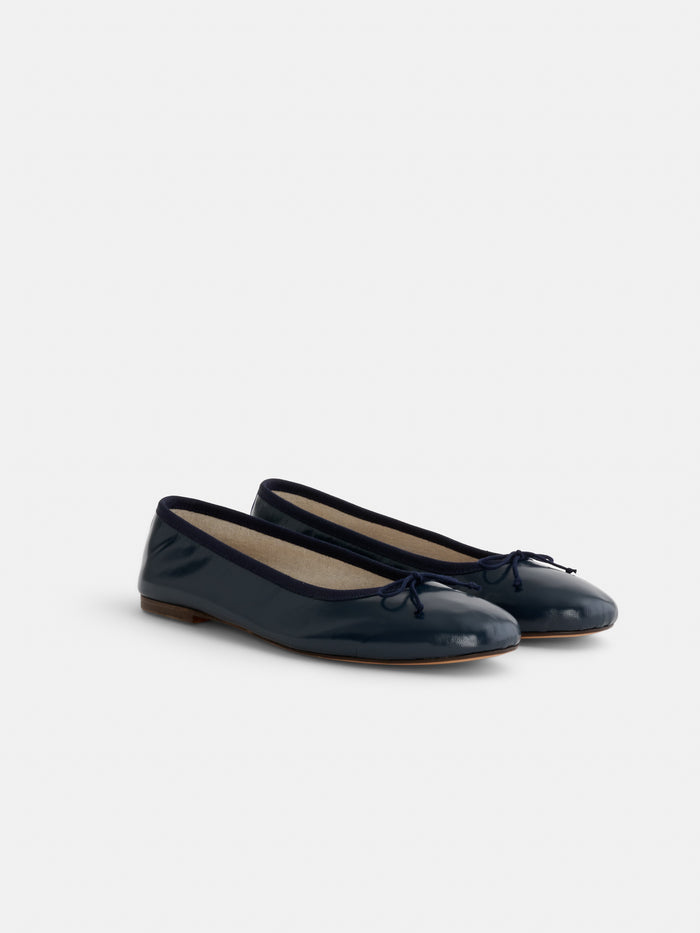 The Prince Street Ballet Flat, handmade in Florence from fine Italian leather, features a shiny dark navy blue finish, rounded toes, small front bows, and flat brown soles. Displayed on a plain light background.