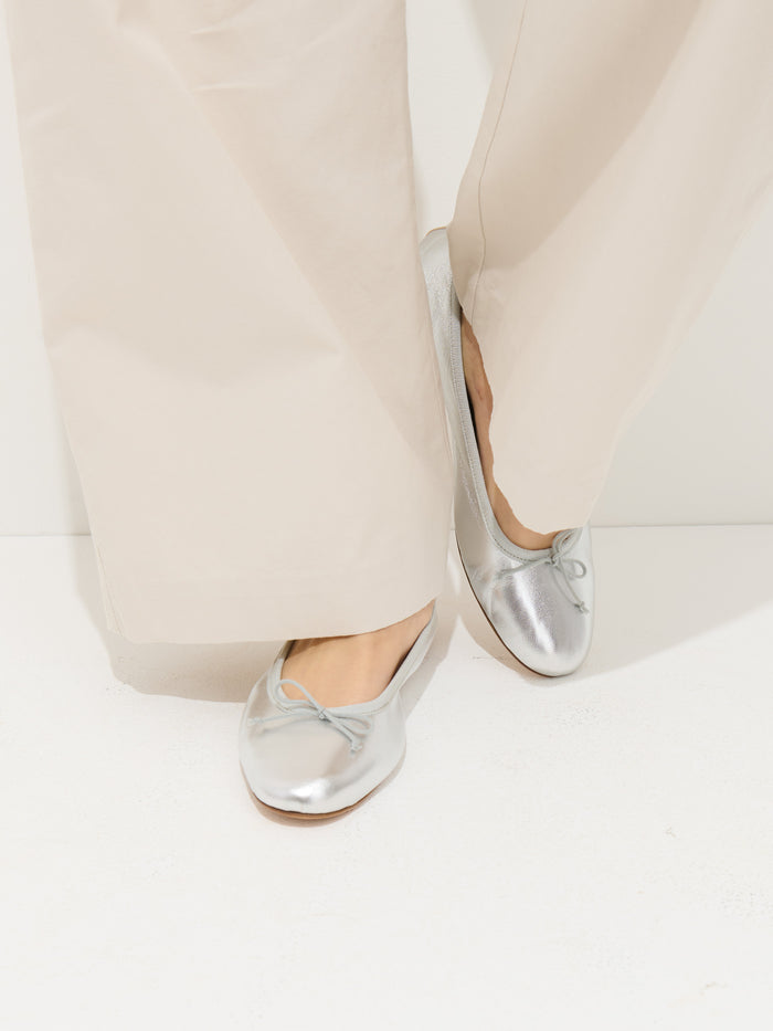 A person wears wide-leg, light beige pants and metallic silver Prince Street Ballet Flats—handmade in Florence with a small bow—while standing on a white surface.