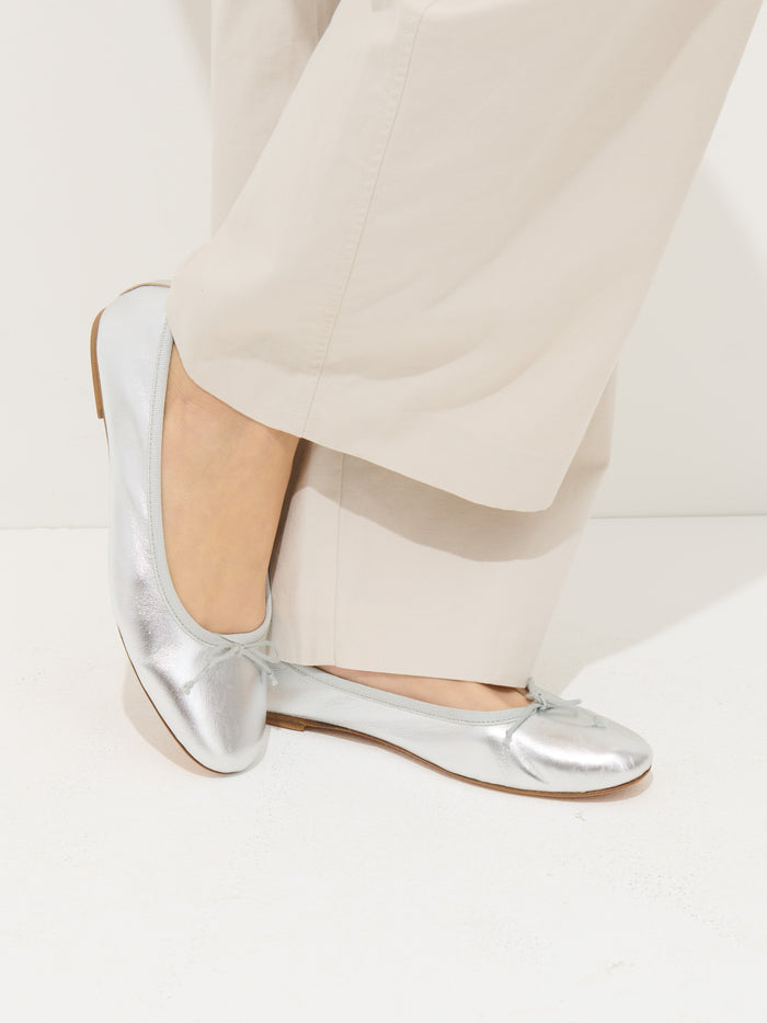 A person in light beige pants wears Prince Street Ballet Flats—metallic silver, handmade in Florence from fine Italian leather—standing on a white surface with one foot crossed over the other.
