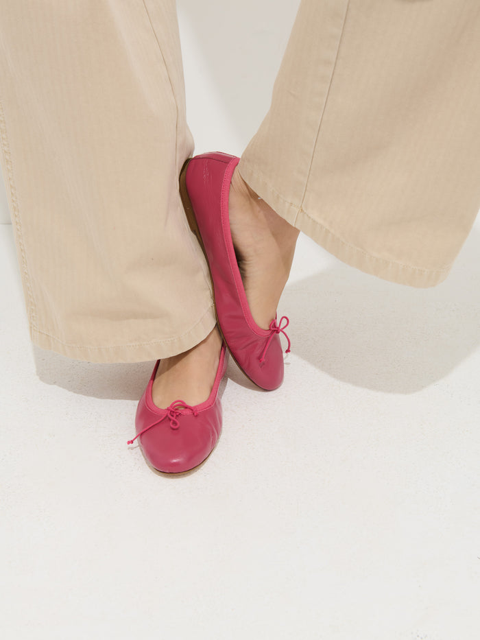 A person wearing beige pants and the Prince Street Ballet Flat in bright pink Italian leather with small bows, standing on a white surface with one foot slightly crossed over the other.