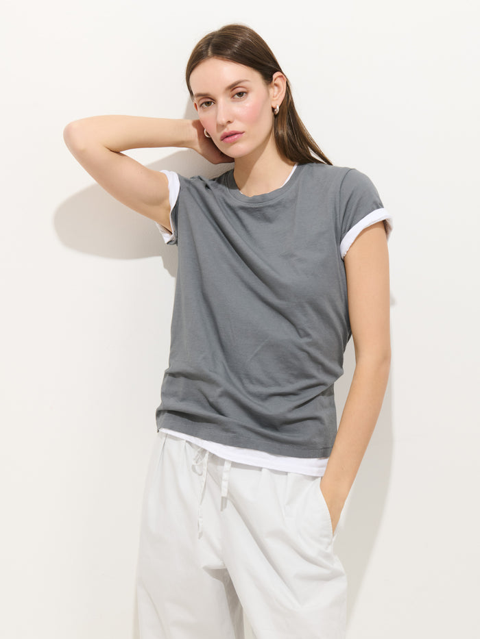 A young woman with long brown hair stands against a white wall, wearing the Prospect Tee In Cotton Jersey over a white shirt and white pants, with one hand in her pocket and the other on her head, calmly looking at the camera.