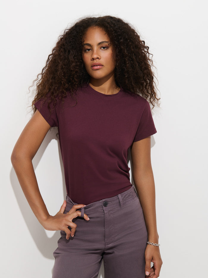 A woman with long curly hair models the Prospect Tee In Cotton Jersey, a maroon garment-dyed t-shirt made in Peru, paired with gray pants. She stands against a plain white wall with one hand on her hip and a neutral expression.