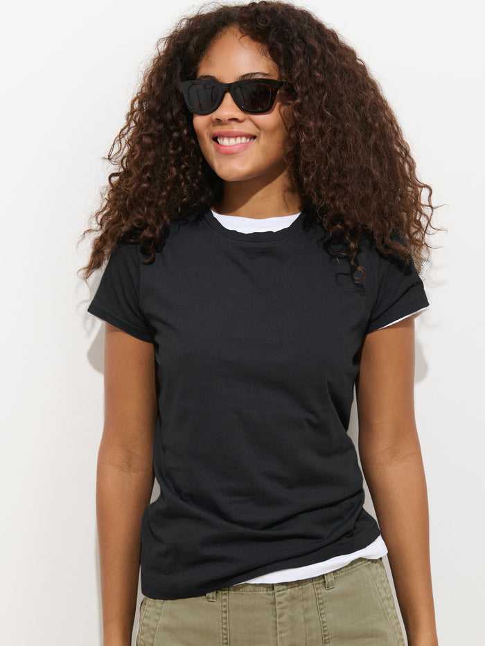 A woman with curly hair, wearing the Prospect Tee In Cotton Jersey over a white shirt and olive-green pants, smiles in sunglasses against a plain white background.