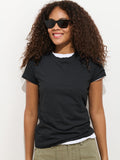 A woman with curly hair, wearing the Prospect Tee In Cotton Jersey over a white shirt and olive-green pants, smiles in sunglasses against a plain white background.