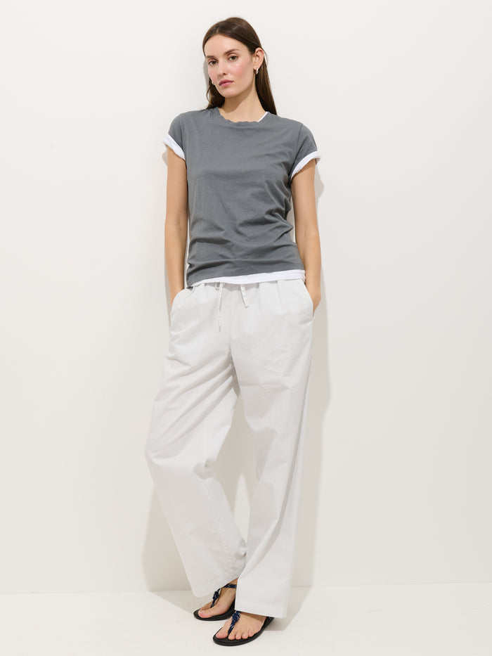 A woman with long brown hair stands against a white background, wearing the Prospect Tee in Cotton Jersey (garment-dyed gray) with rolled sleeves, loose white pants, and black flip-flops. Her hands rest casually in her pockets.