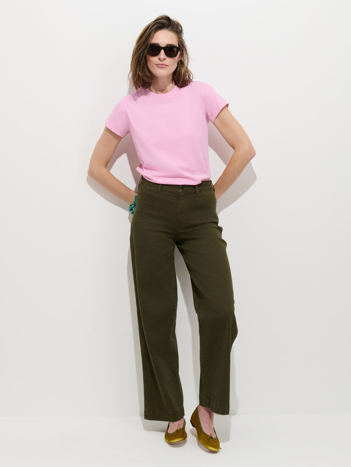 A woman wearing the Prospect Tee In Cotton Jersey in pink, paired with olive green wide-leg pants and yellow shoes, stands against a white background with her hands in her pockets and sunglasses on.