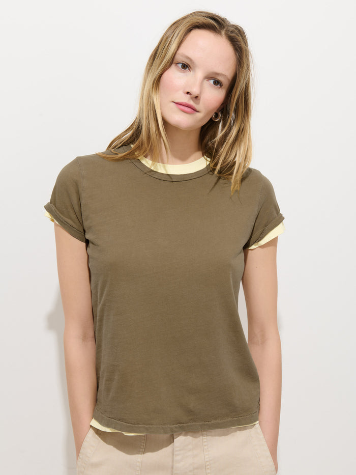 A woman with straight, blonde hair stands against a white background, wearing the Prospect Tee In Cotton Jersey—an olive green, 100% cotton, garment-dyed T-shirt with light yellow trim—and beige pants. She looks slightly to the side, expression neutral.