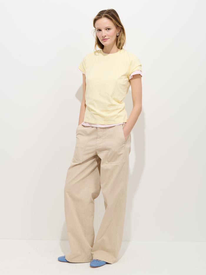 A person stands against a plain white background, wearing the Prospect Tee in Cotton Jersey, light yellow and made in Peru, paired with beige wide-leg pants and blue shoes. One hand is in their pocket and they have a relaxed expression.