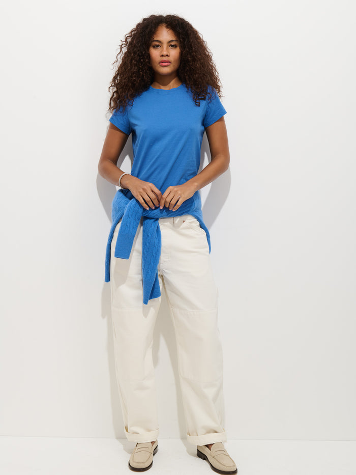 A woman with curly hair stands against a white background, wearing the Prospect Tee in Cotton Jersey (blue), cream wide-leg pants, beige shoes, and a garment-dyed blue cotton sweater tied around her waist.