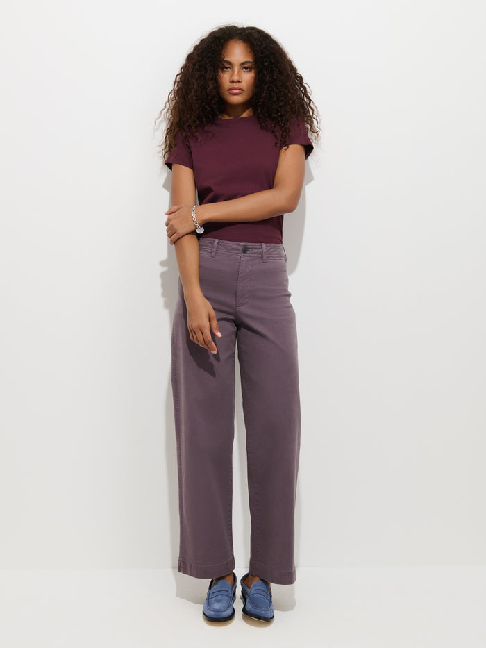 A woman with curly hair wears the Prospect Tee In Cotton Jersey in maroon, paired with wide-legged purple pants and blue shoes. She stands against a white background with her arms loosely crossed.