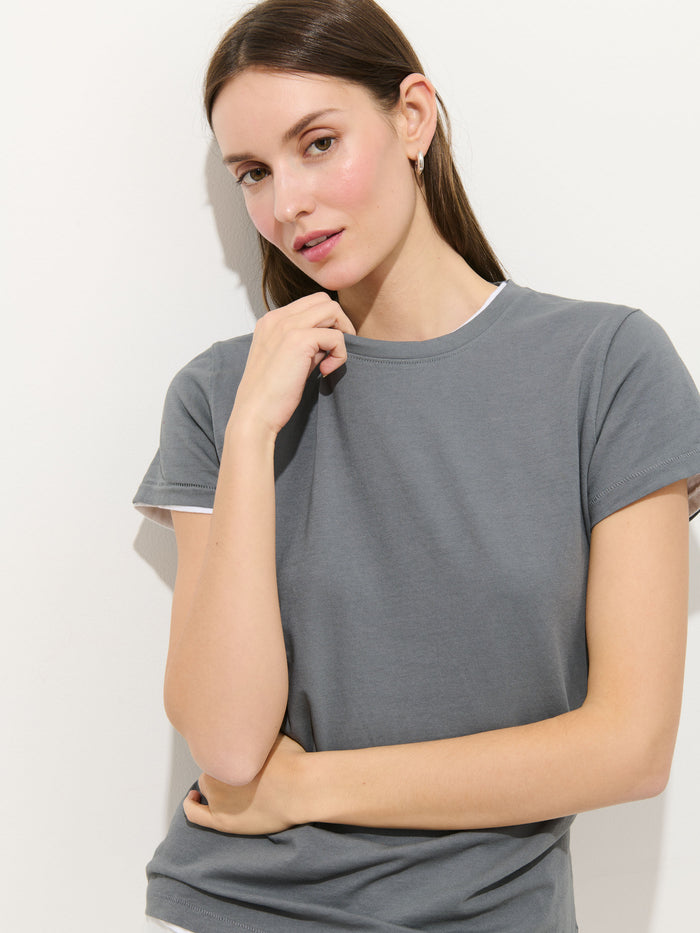 A woman with long brown hair wears the Prospect Tee in cotton jersey—gray, garment-dyed, 100% cotton, made in Peru—as she poses against a plain white background, resting one arm across her body and touching her chin.