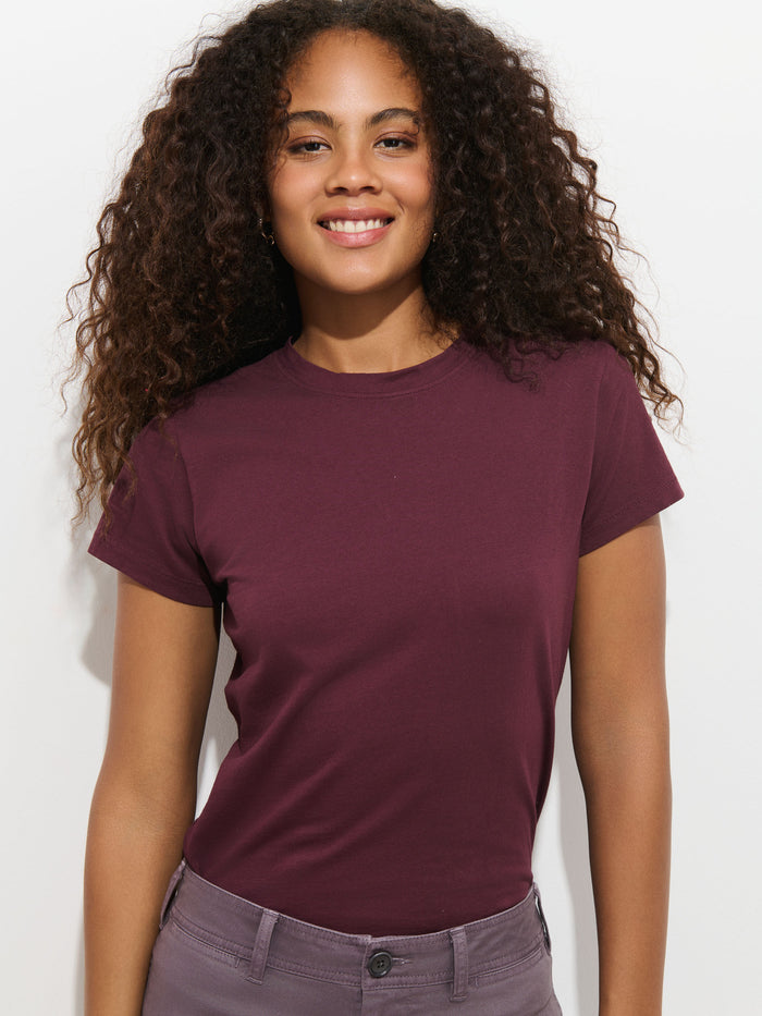 A woman with long, curly hair smiles at the camera. She is wearing the Prospect Tee in Cotton Jersey, a short-sleeved, burgundy t-shirt made in Peru, along with gray pants, standing against a plain white background.