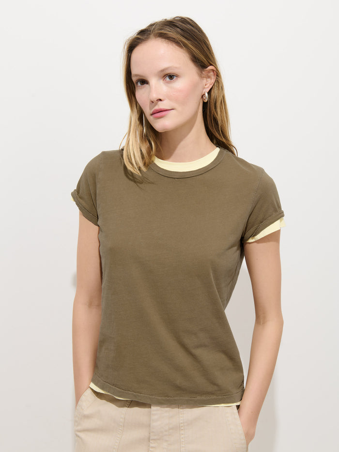 A woman with light brown hair wears the olive green Prospect Tee in Cotton Jersey over a light yellow top and beige pants, standing against a plain white background with her hands in her pockets, looking slightly to the side.