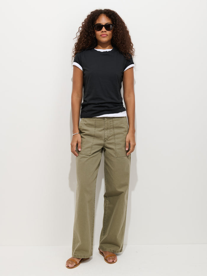 A person with curly hair and sunglasses stands against a white background, wearing the Prospect Tee In Cotton Jersey in garment-dyed black over a white shirt, paired with khaki wide-leg pants and brown sandals.