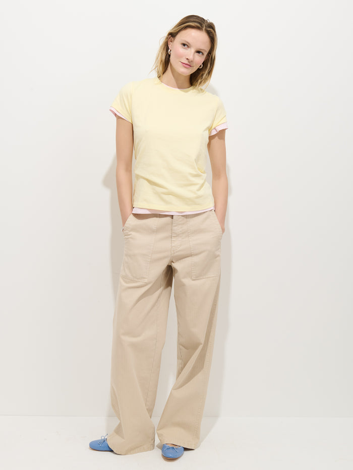 A woman stands against a white background, wearing the Prospect Tee In Cotton Jersey in pale yellow, beige wide-leg pants with large pockets, and light blue shoes. She has her hands in her pockets and is smiling slightly.