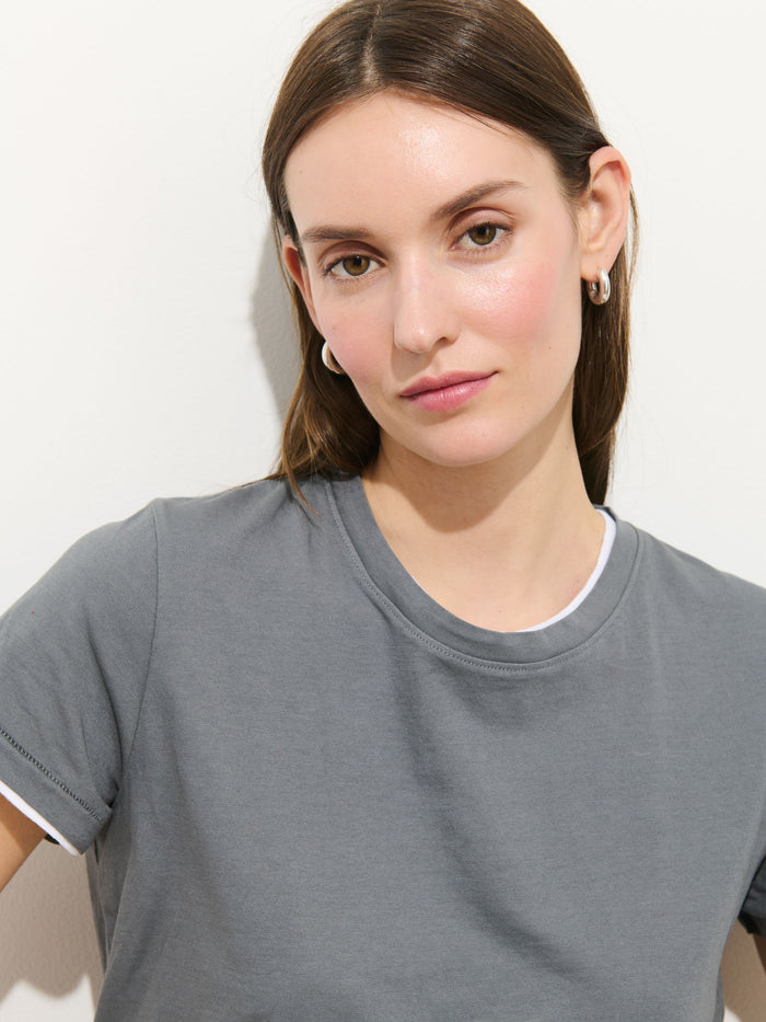 A woman with straight brown hair wears the grey Prospect Tee In Cotton Jersey and silver hoop earrings, looking at the camera with a neutral expression against a plain white background.