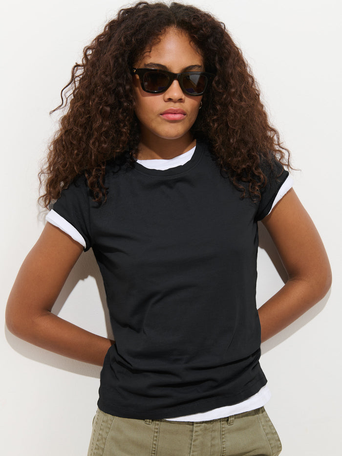 Wearing sunglasses and olive green pants, a person with long curly hair models the Prospect Tee In Cotton Jersey—a garment-dyed black t-shirt with white sleeve cuffs—standing hands-on-hips against a plain white background.