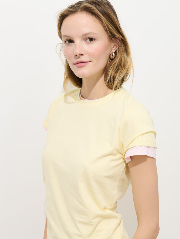 A woman with light brown hair and fair skin stands against a plain white background, wearing the Prospect Tee in Cotton Jersey, a pale yellow garment-dyed shirt made in Peru with light pink sleeve lining. She smiles slightly at the camera.