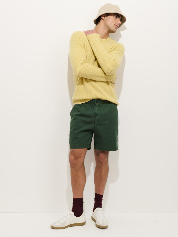 A man in a beige bucket hat, yellow sweater, Pull On Short In Cotton Herringbone, burgundy socks, and white sneakers stands against a plain white background with his arms crossed over his chest.