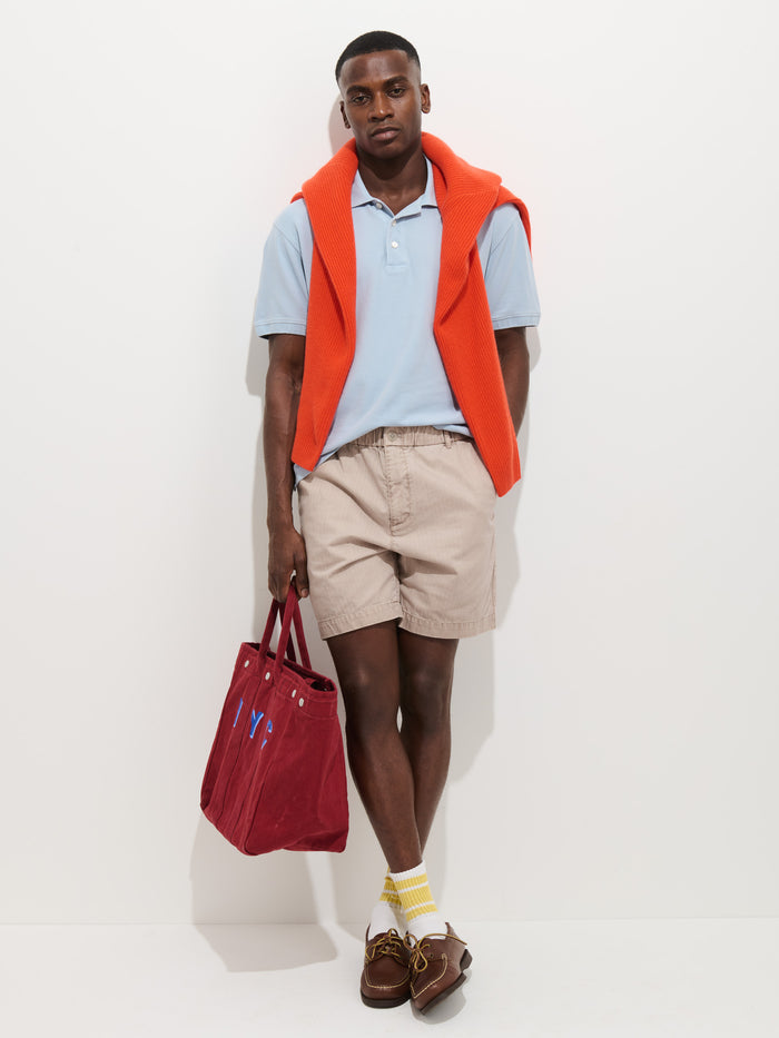 A man stands against a white wall wearing a light blue polo, Pull On Short In Cotton Herringbone with drawstring waist, brown boat shoes, yellow socks, an orange sweater draped over his shoulders, holding a red tote bag and looking at the camera.