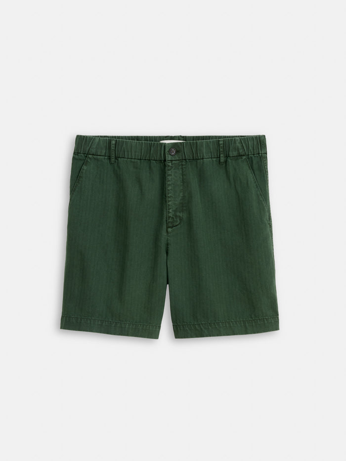 The Pull On Short In Cotton Herringbone features a dark green hue, elastic waistband, front button closure, and side pockets, shown on a plain white background.