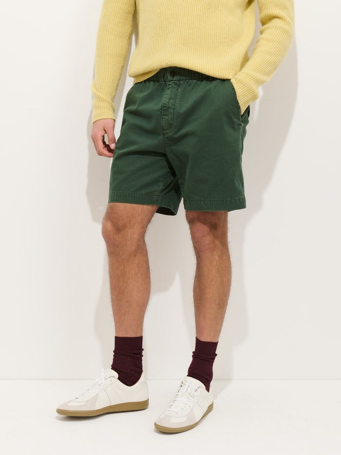 A person stands against a white background with hands in pockets, wearing a light yellow sweater, Pull On Short In Cotton Herringbone, burgundy socks, and white sneakers.