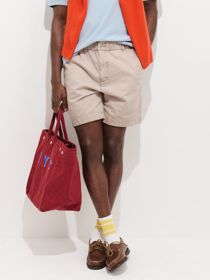 A person wearing Pull On Short In Cotton Herringbone (beige, drawstring waist), a light blue shirt, orange sweater over their shoulders, yellow and white striped socks, brown boat shoes, and holding a red tote bag stands against a white background.