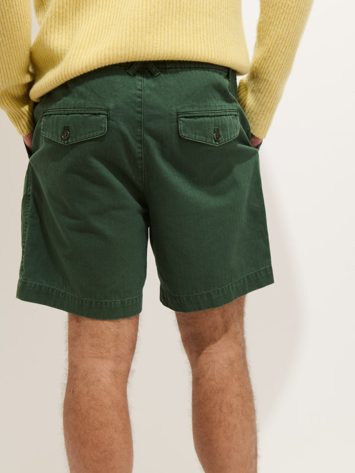 A person wearing a yellow sweater and Pull On Short In Cotton Herringbone stands with their back to the camera, hands in pockets. The shorts are green with buttoned back pockets.