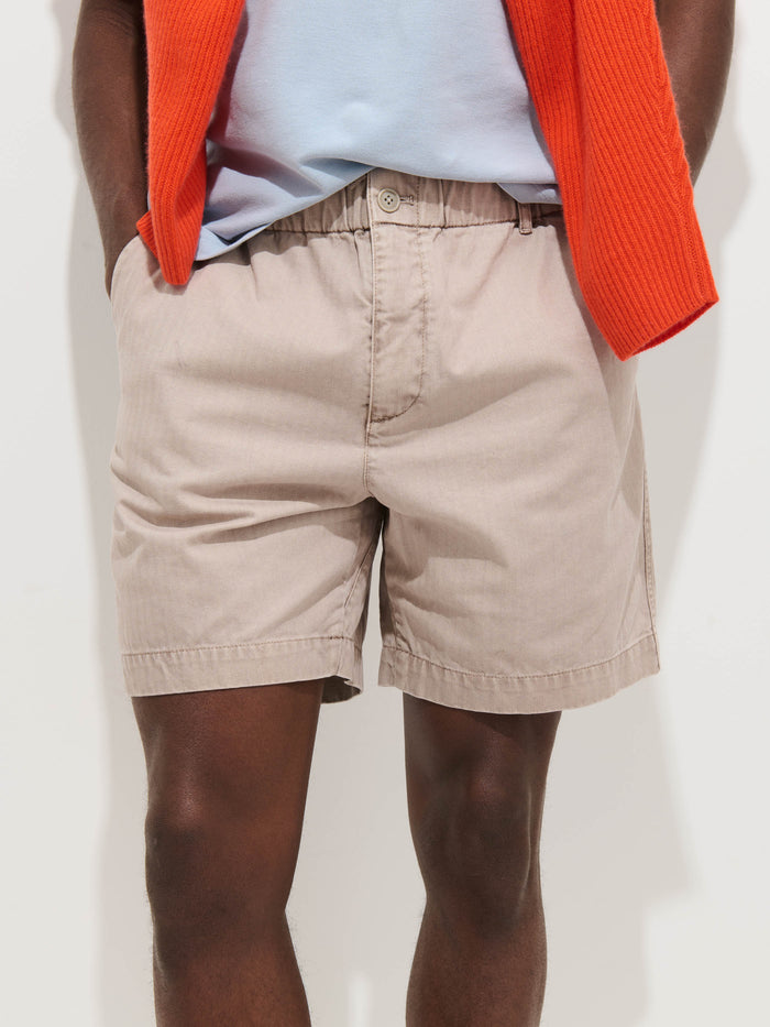 A person wears the Pull On Short In Cotton Herringbone in light beige with a drawstring waist, paired with a pale blue shirt and an orange sweater draped over their shoulders, standing against a plain white background.