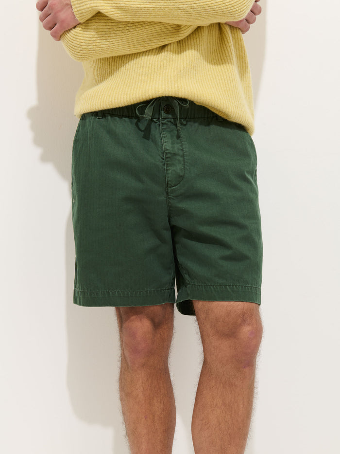 A person stands against a light background, wearing a yellow sweater and green Pull On Short In Cotton Herringbone. Their arms are crossed, and their face is not visible in the image.
