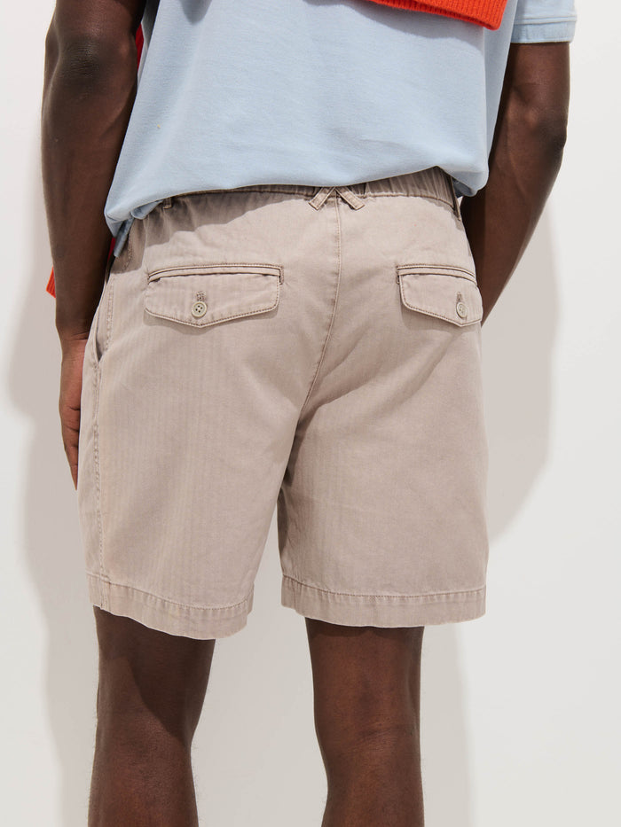A person is shown from the waist down wearing Pull On Short In Cotton Herringbone, light beige with buttoned back pockets. A light blue shirt is tucked in and a red item is draped over one shoulder against a plain white background.