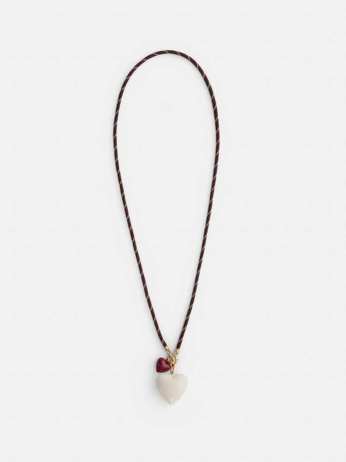 The Roxanne Assoulin Long Happy Cord Charm Necklace features a puffy white heart-shaped pendant, a smaller red heart charm near the clasp, and colorful dark brown cording.