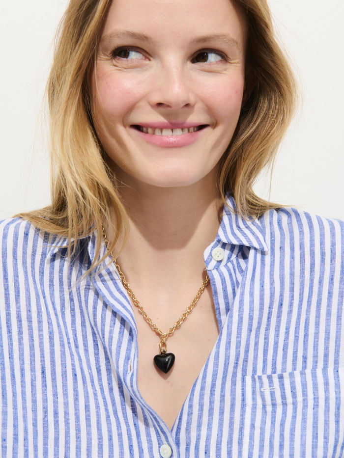 A smiling woman in a blue and white striped shirt wears the Roxanne Assoulin Mini Puffy Heart Necklace, its gold and black puffy heart pendant adding a charming touch as she looks slightly to the side against a plain background.