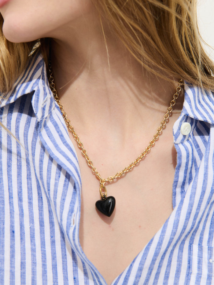 A woman wears the Roxanne Assoulin Mini Puffy Heart Necklace, featuring a gold brass cable chain and black puffy heart pendant, along with a blue and white striped button-up shirt. The image is cropped to show her neck and part of her face.